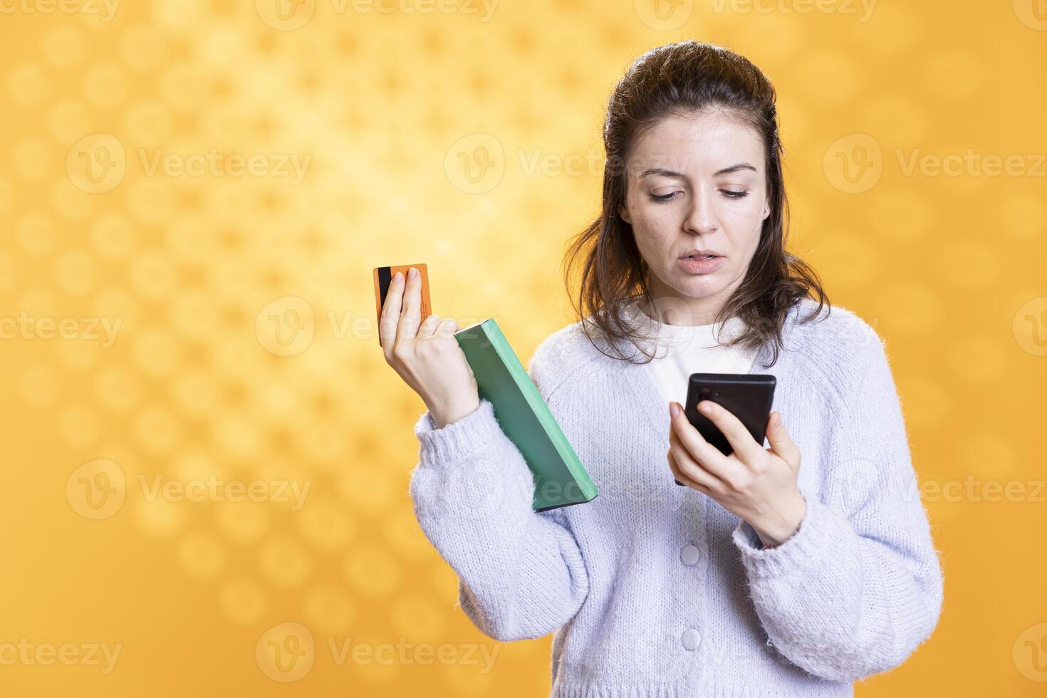 Woman adding credit card information on smartphone while holding book, studio background. Person with academic textbook in hand purchasing more educational material using phone photo