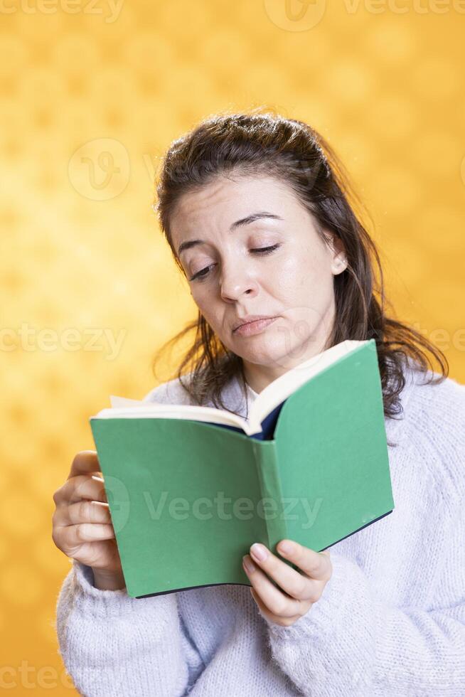 Woman feeling unimpressed while reading bad book, enjoying hobby during leisure time. Bookaholic person with dull novel in hands disappointed by boring story, isolated over studio background photo