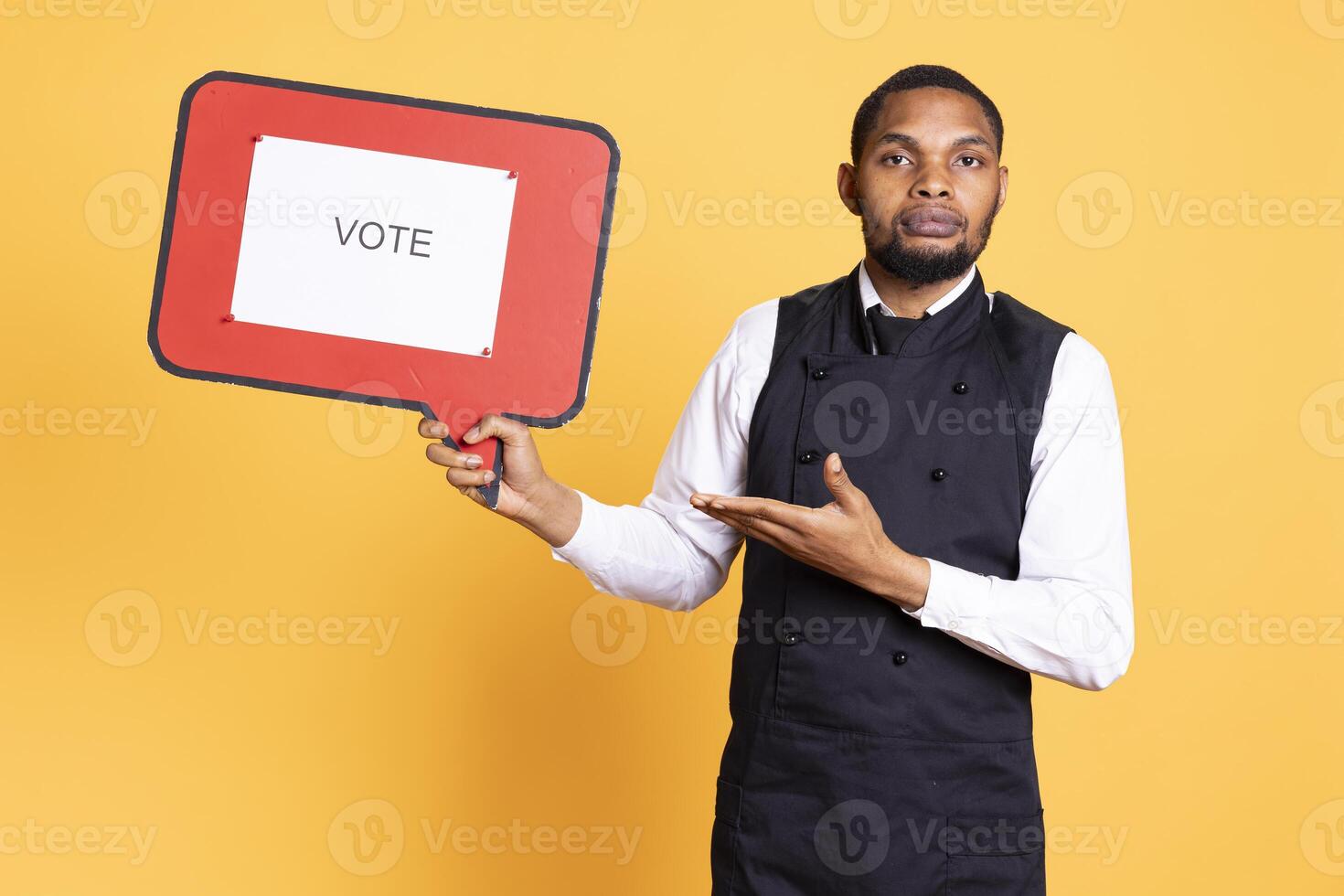 Waiter does an advertisement for politics and showing a speech bubble with the message to vote, standing against yellow background. Restaurant staff holds a cardboard sign. photo