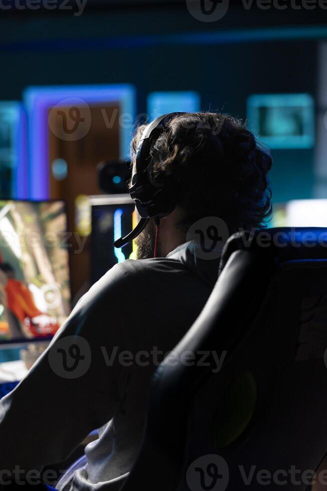 Man relaxing by playing entertaining videogame on PC at computer desk late at night. Close up shot of gamer wearing headphones, shooting enemies in online multiplayer first person shooter photo