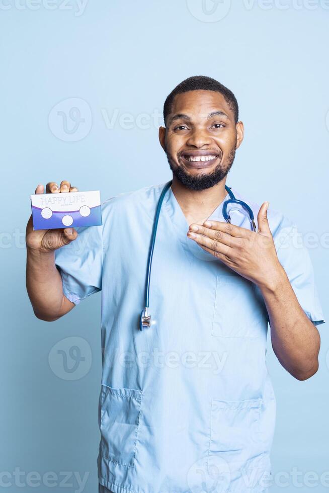 Portrait of doctor in scrubs recommending vitamins and supplements for wellness, holding medicine box for patients in studio. Hospital employee pointing at tablets for chronic disease. photo