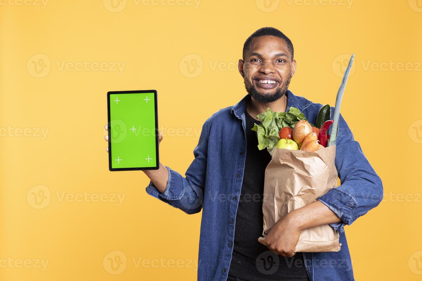 African american smiling adult presenting mockup display on a tablet, carrying bag of groceries in studio. Relaxed cheerful guy showing isolated greenscreen layout on his gadget. photo