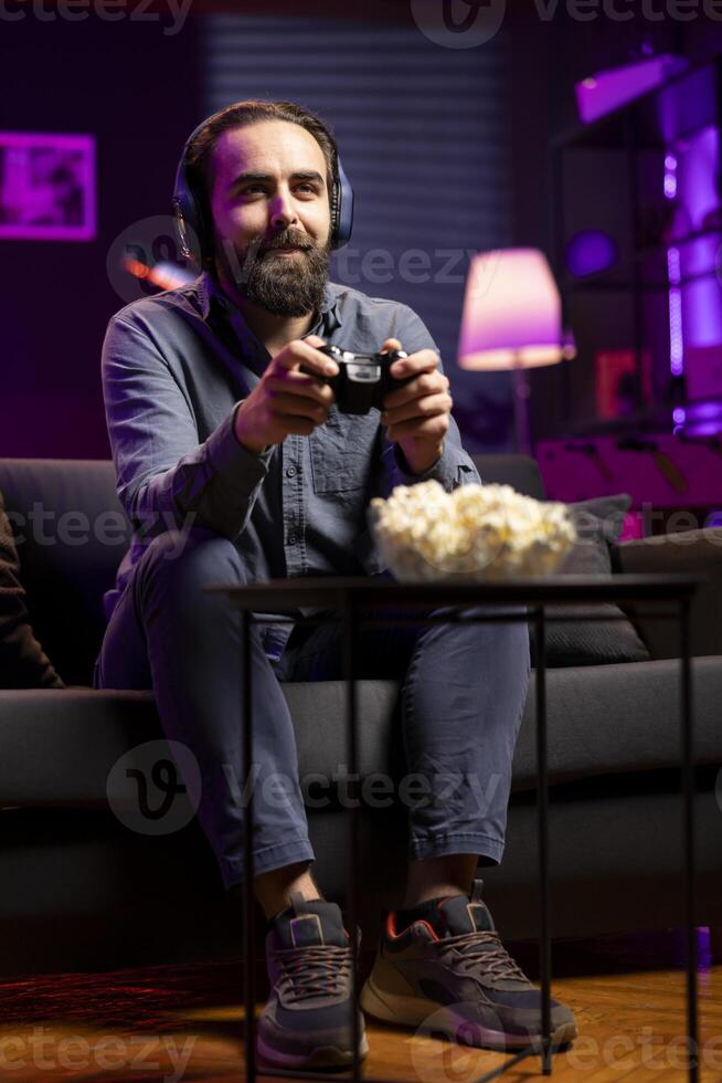 Smiling man showing positive emotions while playing videogames on gaming console with teammates. Happy player enjoying defeating foes in game, discussing with friends through headset photo