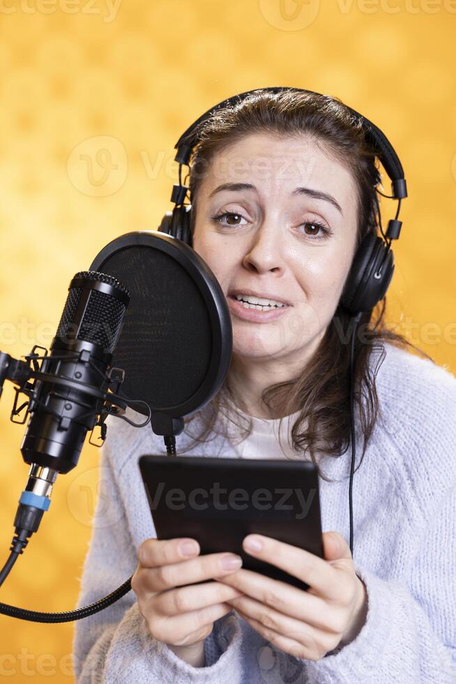 Portrait of smiling woman reading ebook from ereader, recording audiobook using microphone, studio background. Jolly narrator using mic to produce digital recording of novel, reading text from tablet photo