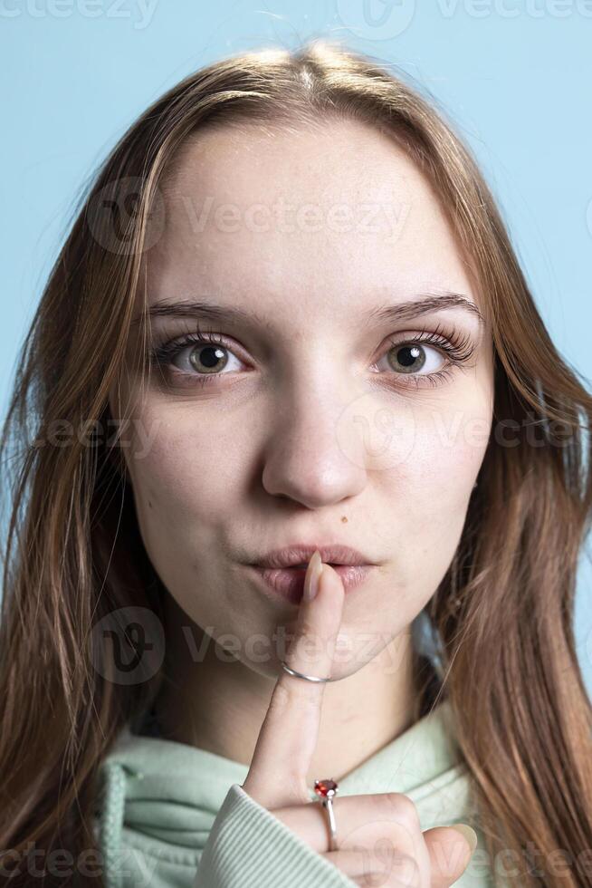 Stern teenager doing shushing hand gesturing, irritated by loud noises. Extreme close up shot of girl placing finger on lips, doing quiet sign gesture, blue studio background photo