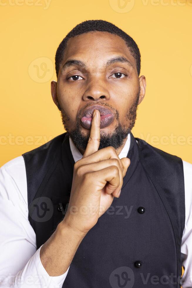 Five star restaurant staff showing the silence symbol and asking for secrecy, presenting the mute sign against yellow background. Waiter in apron asking to keep secret and be quiet. photo