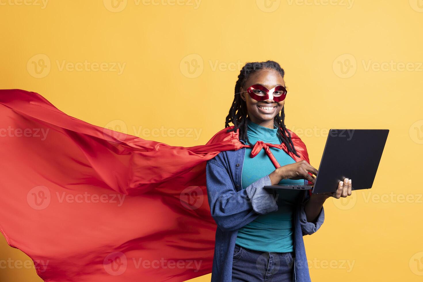Smiling girl wearing superhero costume and mask for Halloween working on laptop, studio background. Portrait of woman dressed as comic book hero hiding identity typing on notebook keyboard photo
