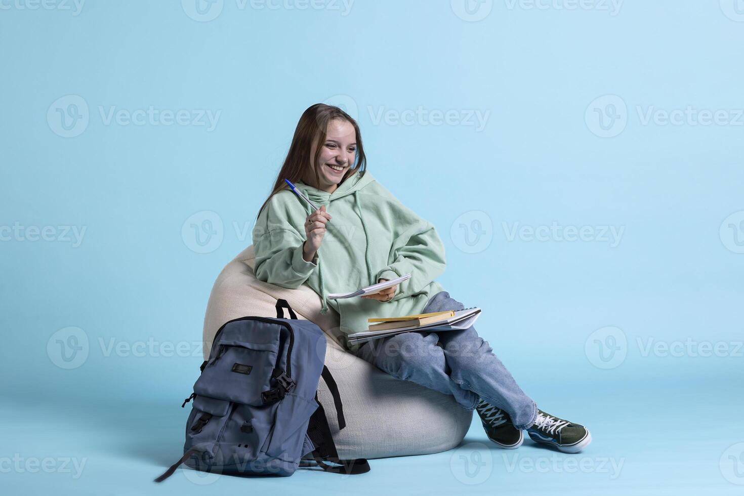 Portrait of jolly girl laughing while sitting on beanbag and writing on notebook, studio background. Overjoyed schoolgirl with notepad in arms feeling amused while preparing homework photo