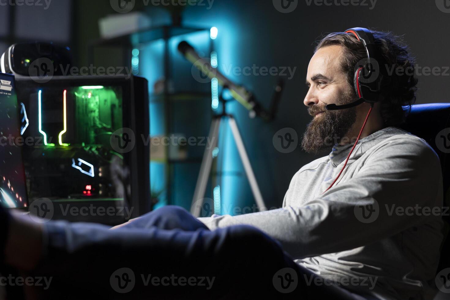 Happy gamer plays online multiplayer videogame with friends, speaking with them using headphones mic. Cheerful player talking with teammates, putting feet up, enjoying leisure time photo