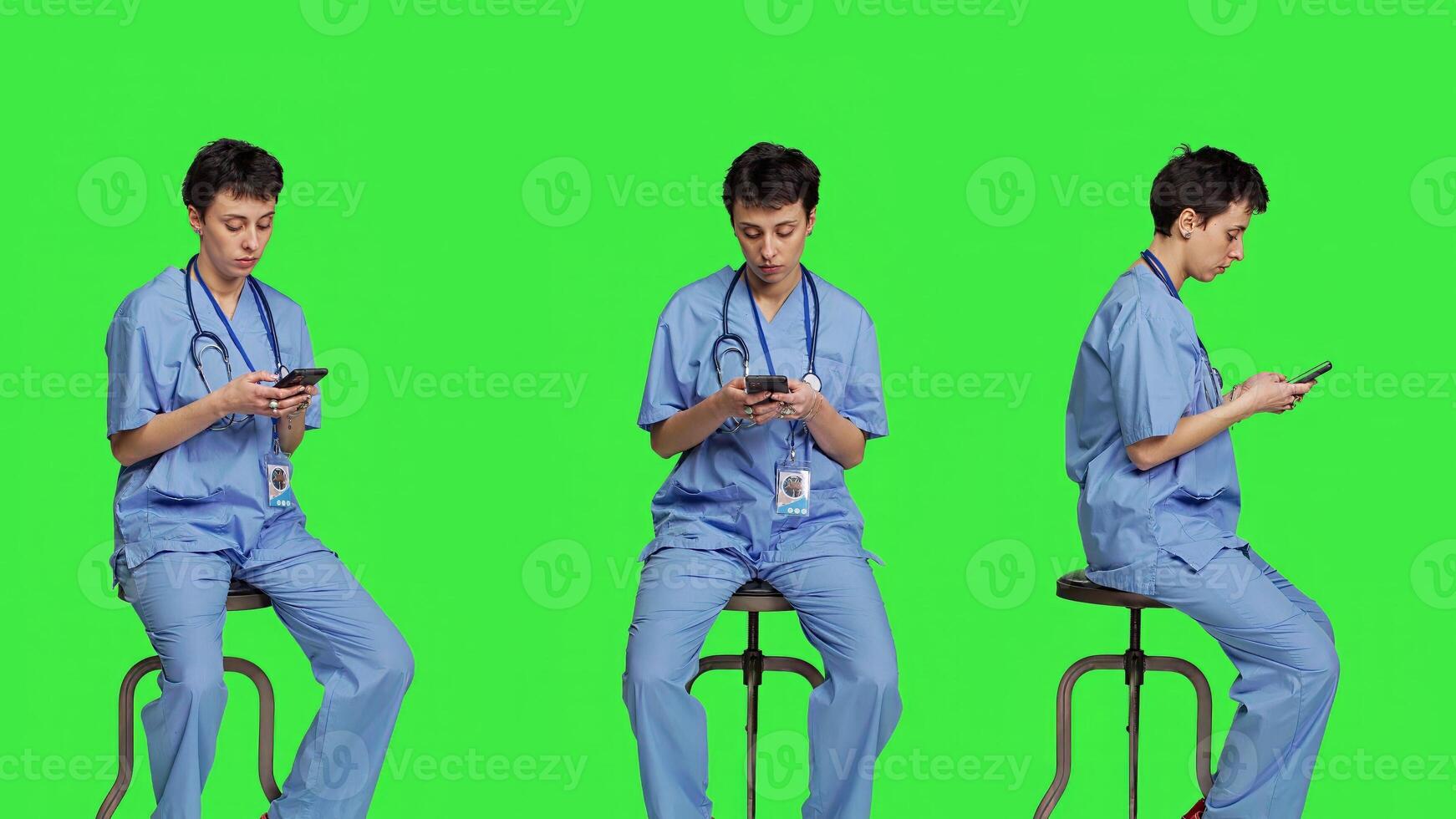 Nurse browsing online websites on smartphone app in studio, sitting on a chair against greenscreen backdrop. Medical specialist in scrubs using mobile phone and social media texting. Camera A. photo