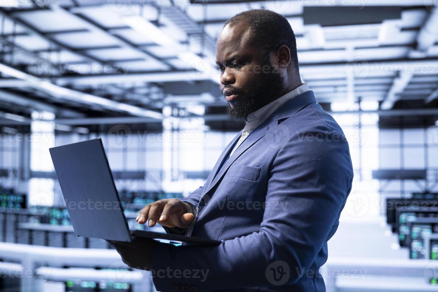 Engineer in server room establishment installing newly ordered storage units using laptop. African american man working in data center facility housing storage rackmounts photo