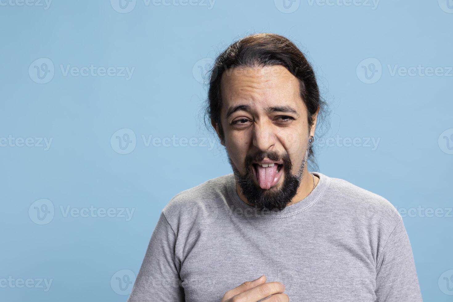 Crazy silly person acting disgusted and unpleasant against blue background, gross discontent guy making a grimace and sticking tongue out. Disgusted annoyed man dissatisfied with something. photo