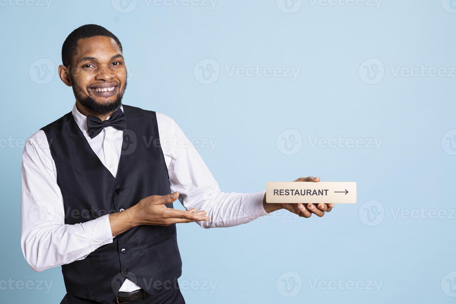 Hotel concierge guiding guests with a wall sign to find all amenities, presenting indicator with the direction to the restaurant against blue background. Confident skilled reception personnel. photo
