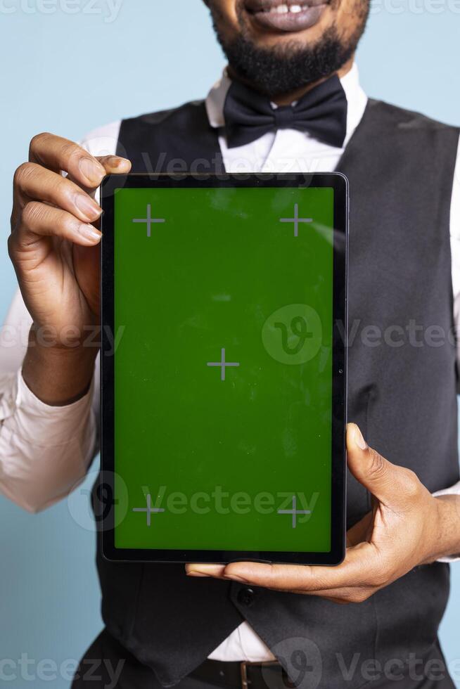 Bellman hotelier showing a tablet with green screen display in studio, presenting mockup chroma key display against blue background. Young hotel concierge working at a resort. photo