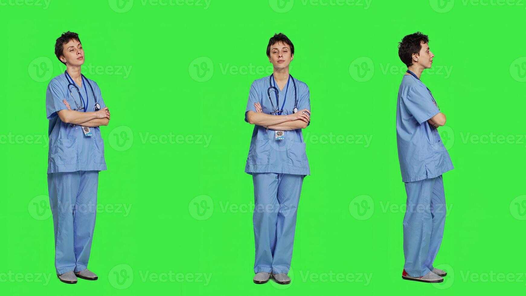 Portrait of smiling medical assistant poses with arms crossed, showing confidence dressed in blue hospital scrubs. Successful nurse standing against greenscreen backdrop, health specialist. Camera A. photo