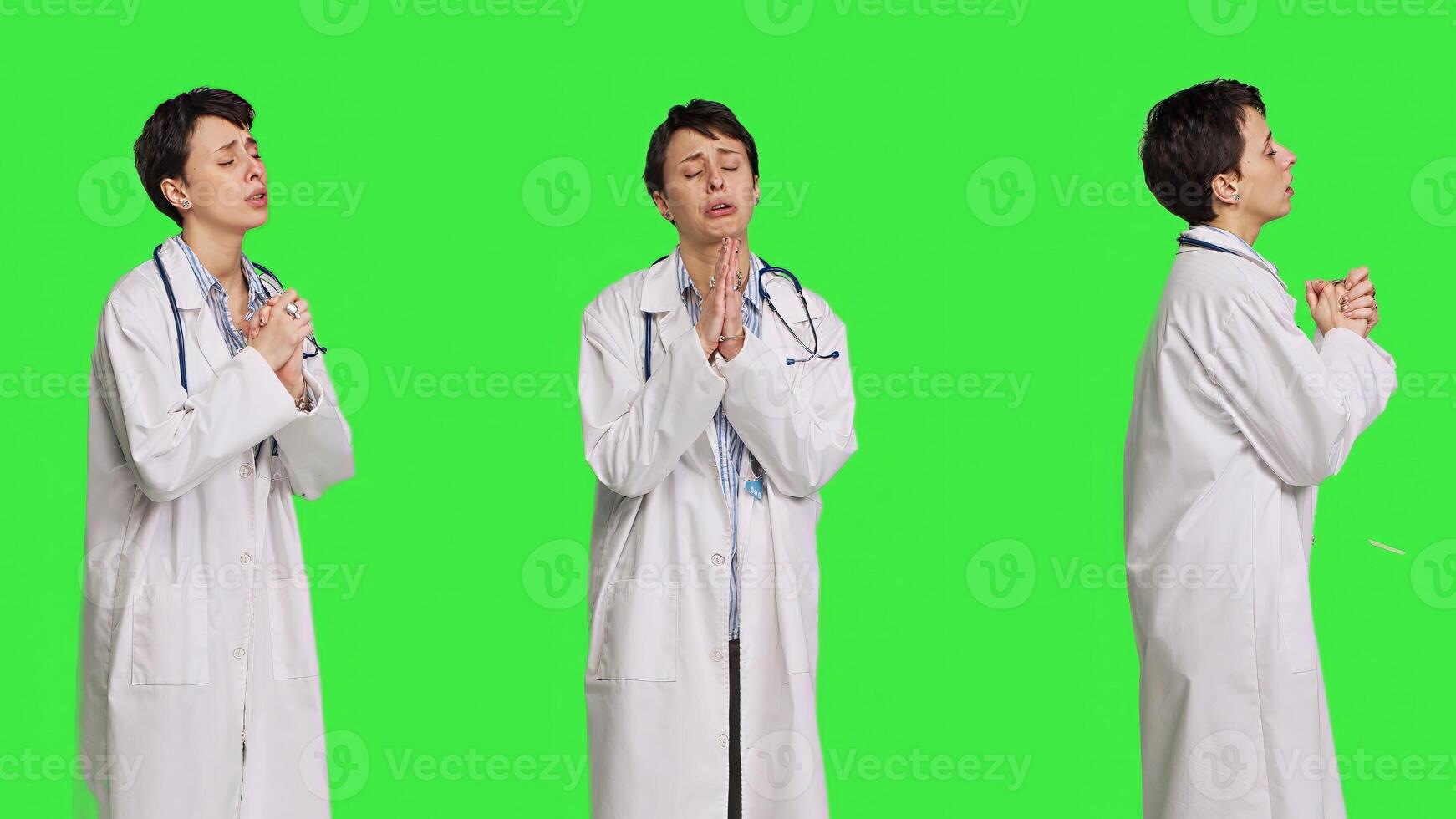 Hopeful physician holding hands in a prayer sign against greenscreen backdrop, asking for peace and forgiveness in studio. Woman medic specialist in white coat does meditation. Camera B. photo