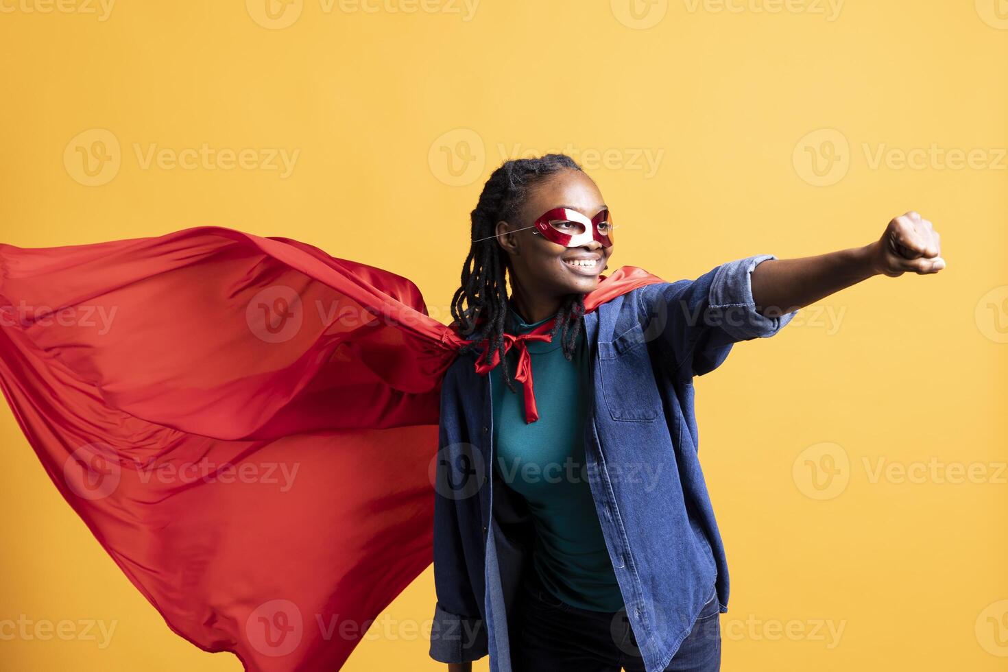 African american girl wearing superhero costume and mask for Halloween doing flying pose, studio background. Woman dressed as comic book hero hiding identity doing heroic hand gesture photo