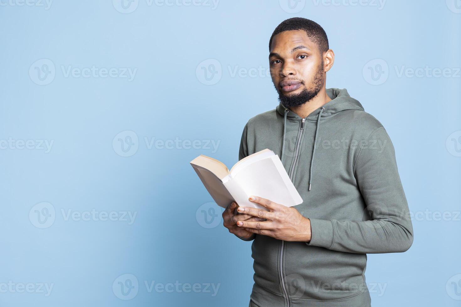 African american cheerful guy enjoys reading a book in studio, feeling happy with literature and novel against blue background. Confident book lover analyzing the synopsis and characters. photo