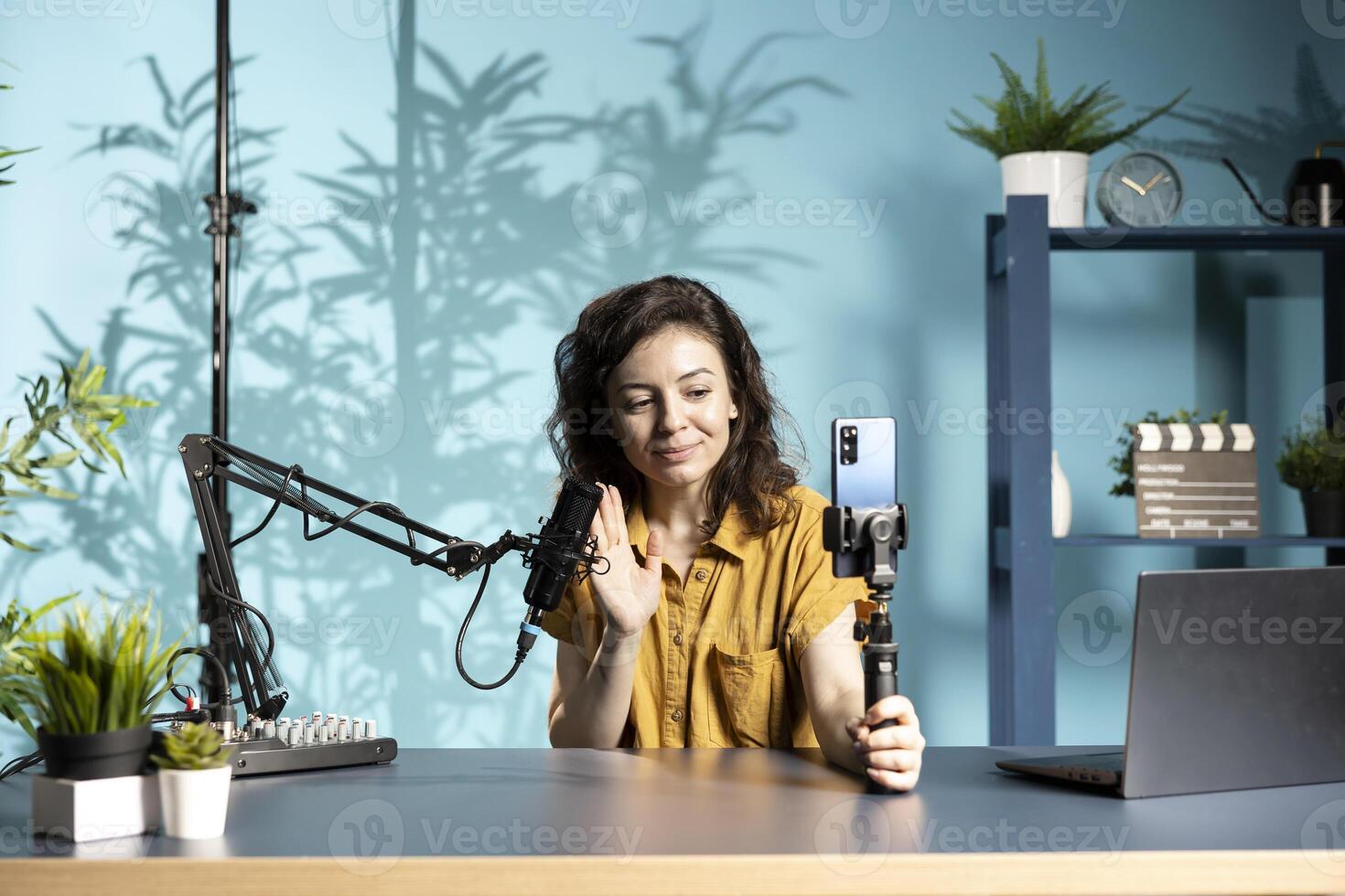 Woman greeting viewers during vlog, using smartphone on selfie stick to capture footage, chatting with online audience. Upbeat content creator saluting fans, filming with mobile phone photo