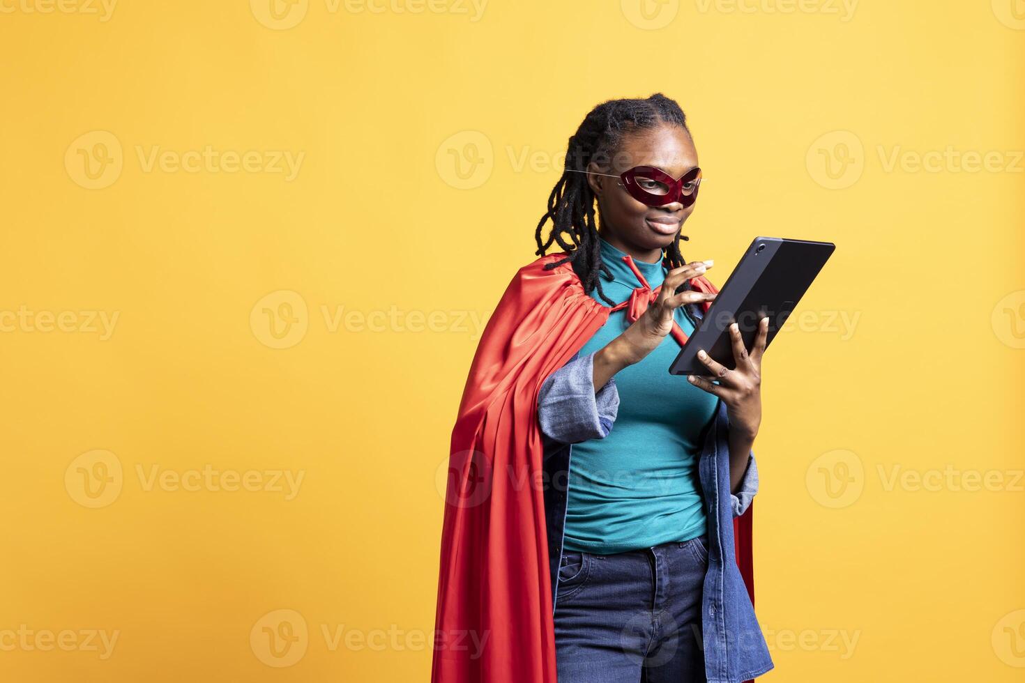 Smiling african american girl portraying superhero hiding identity, working on laptop, studio background. Jolly woman posing as hero in costume, solving tasks on notebook, covering face with mask photo