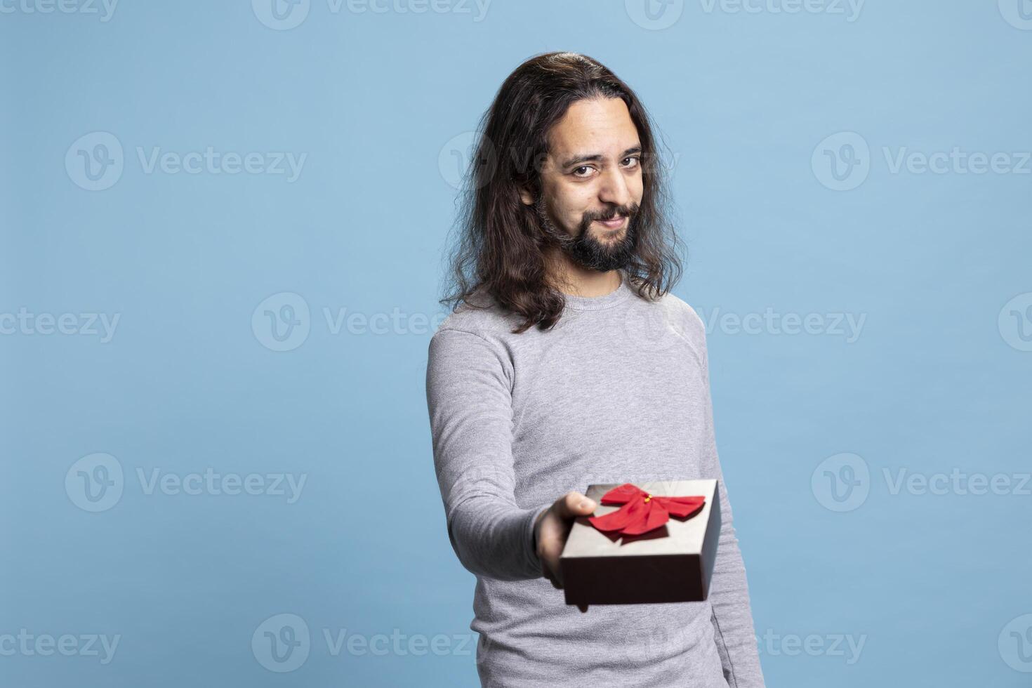 Sweet affectionate guy offering a gift in a cute box with ribbon on camera, giving presents for Christmas or anniversary against blue background. Person in casual wear showing gratitude and love. photo