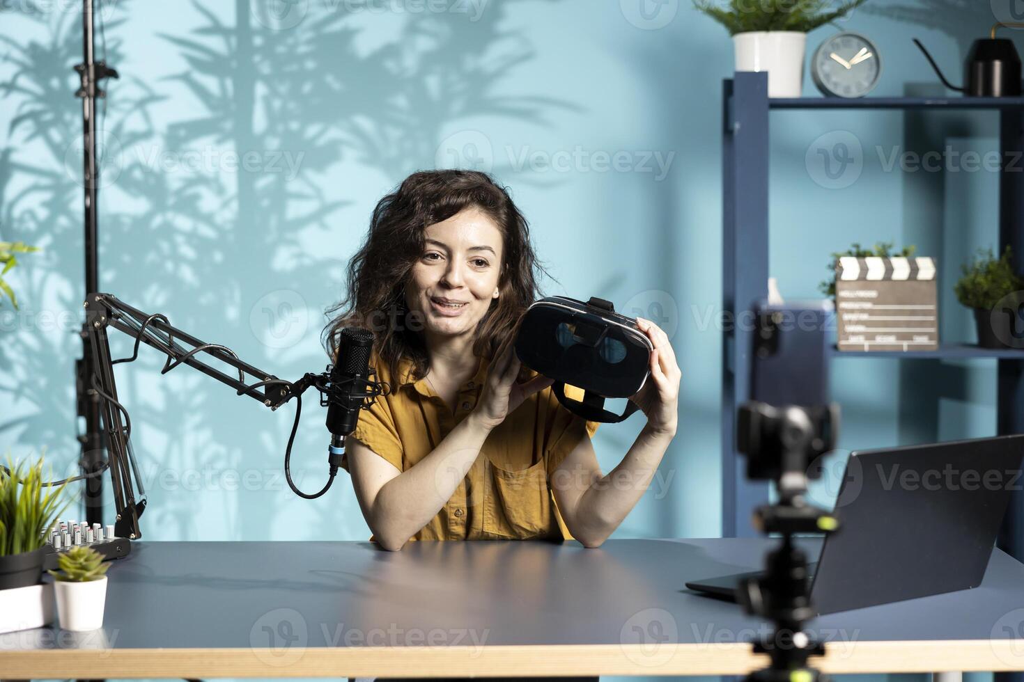 Smiling tech vlogger reviewing VR goggles in living room studio. Happy woman filming virtual reality headset review vlog for gadget enthusiasts using mobile phone and mic photo