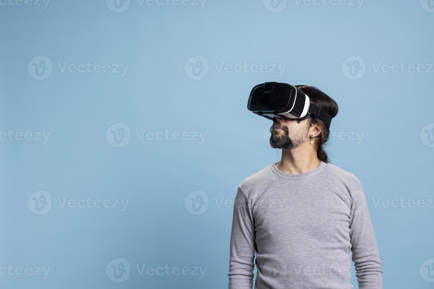 Trendy middle eastern guy using virtual reality headset on camera, using 3D artificial intelligence tech and cyberspace against blue background. Young person does gaming on a vr glasses interface. photo