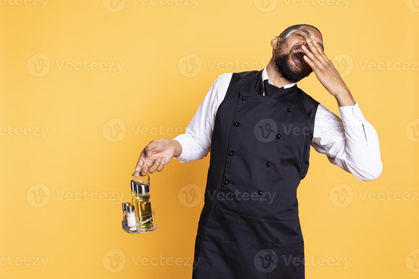 African american waiter feeling very tired in catering and serving industry, yawning and falling asleep while he stands against yellow background. Butler deals with burnout and workload. photo