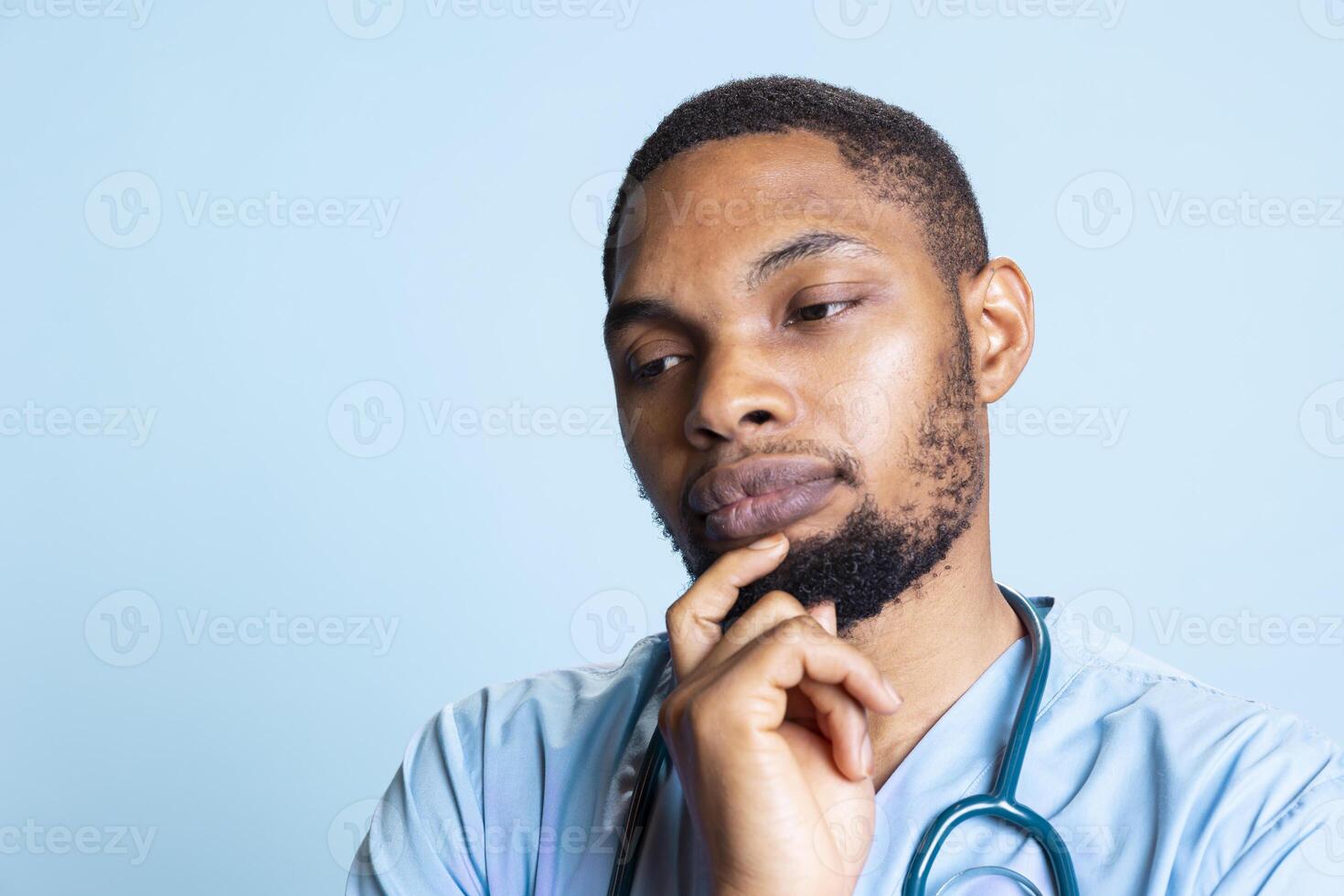 Male medical assistant acting pensive against blue background, thinking about treatment and dosage to cure a patient. Young nurse pondering decision to provide a solution in studio. photo