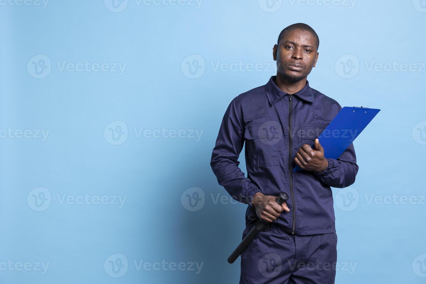 Confident security agent posing with his baton and clipboard files against blue background, using papers to keep track of all responsibilities on the job. Guard protecting the area. photo