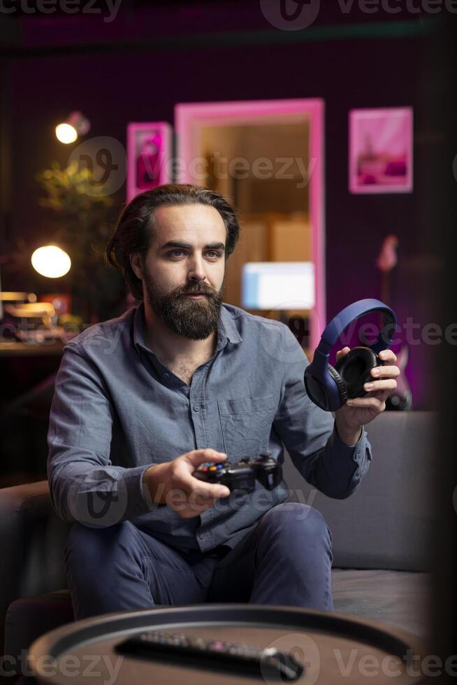 Gamer putting headphones on to chat with friends, sitting down on couch, excited to play games and enjoy leisure time. Man in apartment entertained by videogame on console, relaxing after work photo