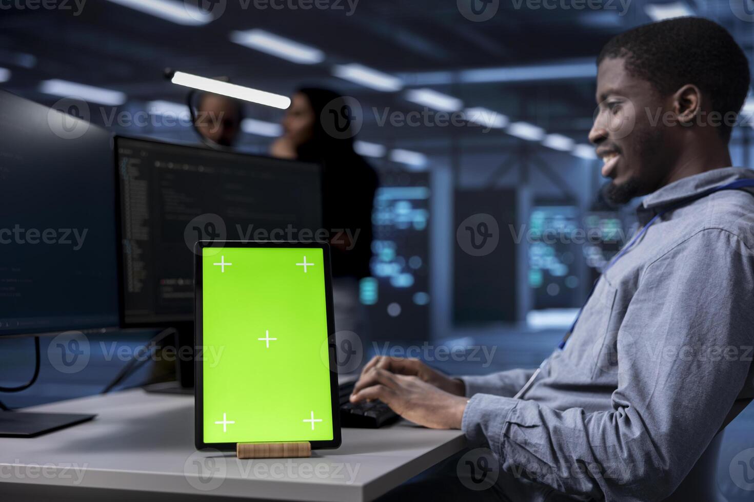 Smiling man working on green screen tablet in modern data center. Happy IT specialist doing IT infrastructure management on server racks in high tech environment using chroma key device photo