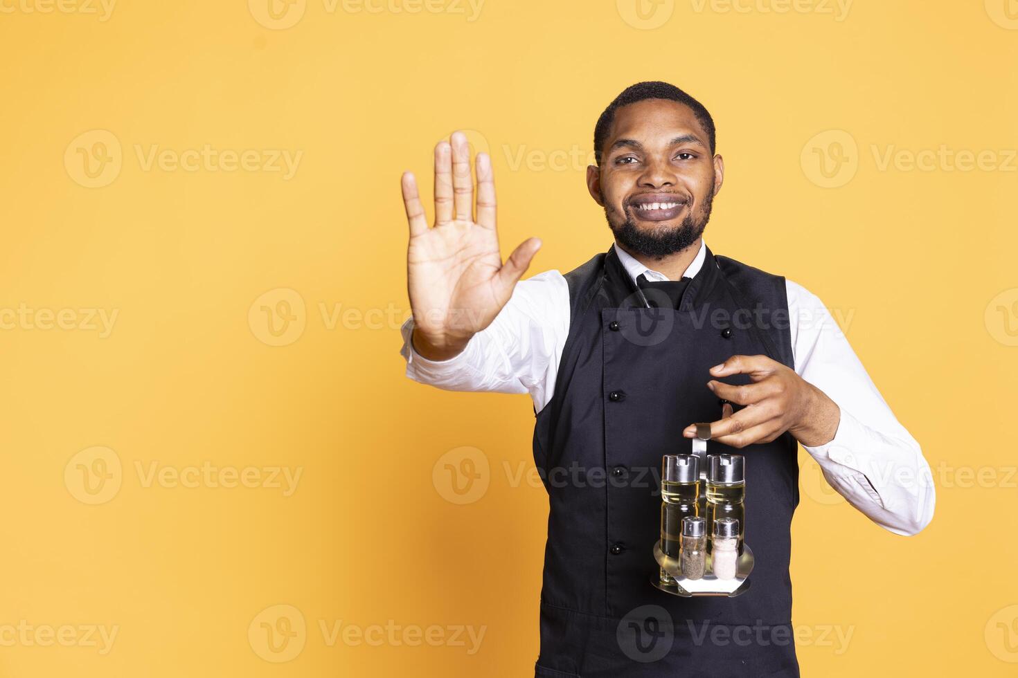 African american butler raising his hand to do a stop sign in studio, expressing his rejection and refusal about something. Restaurant employee waiter carries a cruet set to serve clients. photo