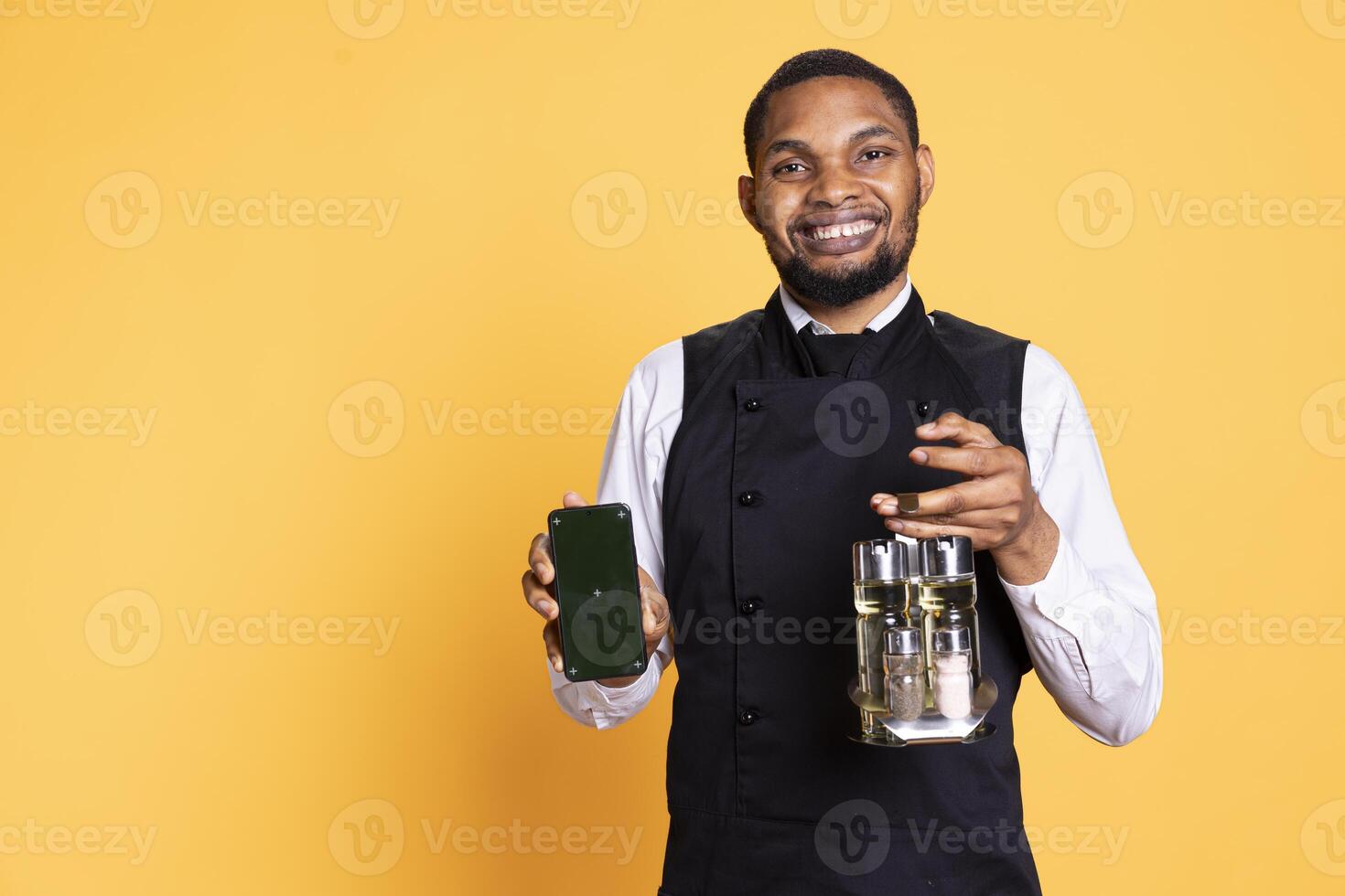 Smiling waiter in uniform presenting a device with isolated chroma key screen, carrying a cruet set to serve clients. Catering industry employee showing mockup display in studio. photo