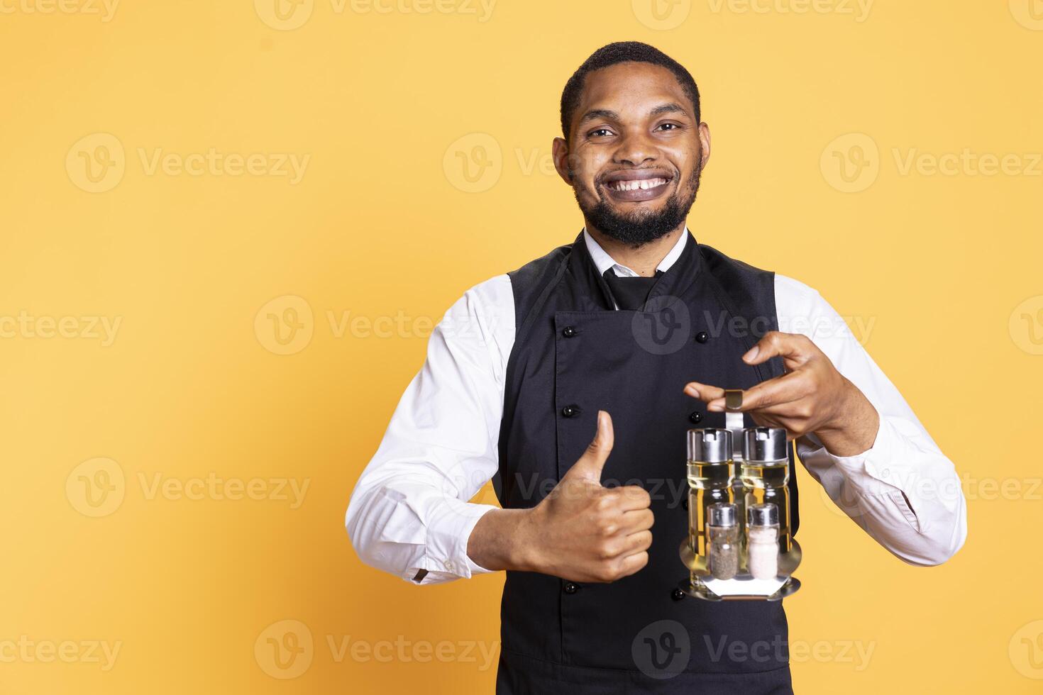 Friendly waiter giving a thumbs up symbol in front of the camera, presenting his agreement and positive emotions in studio. Young staff gives a like and carries the cruet set for tables. photo
