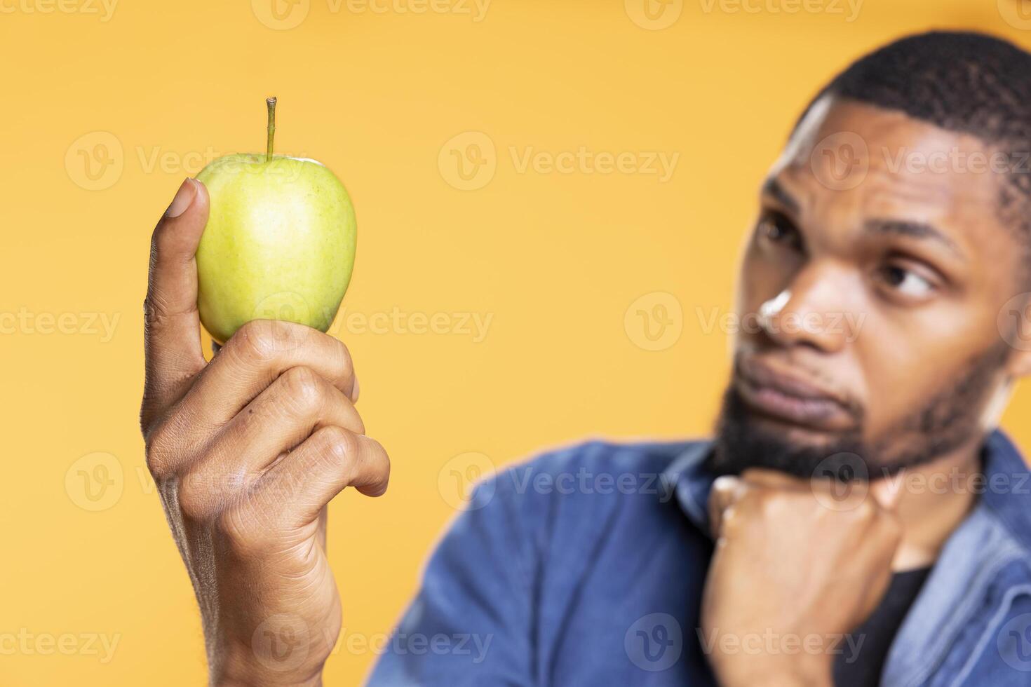 Young person holds a green apple and thinks about a dessert recipe to make with it, preparing to cook something with his ethically sourced organic produce. Pensive guy in front of camera. photo