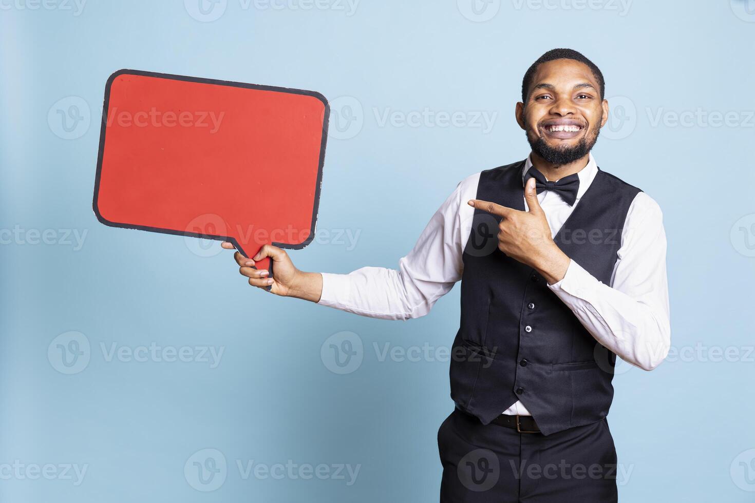 Hotel porter holding a red cardboard speech bubble in front of the camera, pointing at empty commercial advertisement for hospitality industry marketing. Doorman with suit and tie. photo