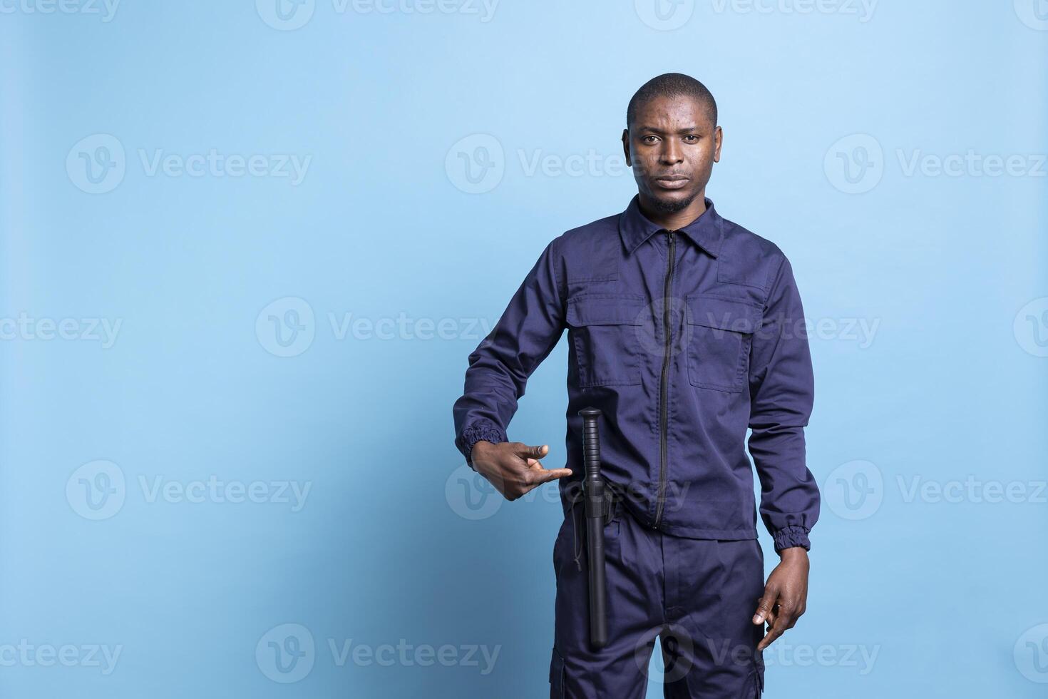 Male security guard pointing to his baton to ensure protection on duty, standing against blue background in patrolling uniform. Confident serious agent presenting his weapon, public safety. photo