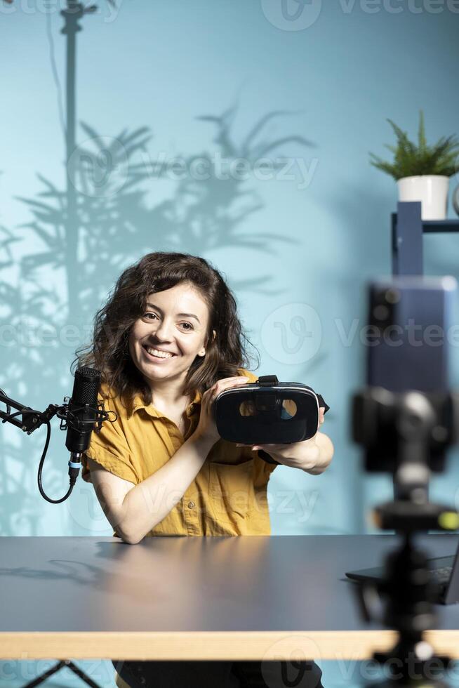 Smiling tech vlogger reviewing VR goggles in living room studio. Happy woman filming virtual reality headset review vlog for gadget enthusiasts using mobile phone and mic photo