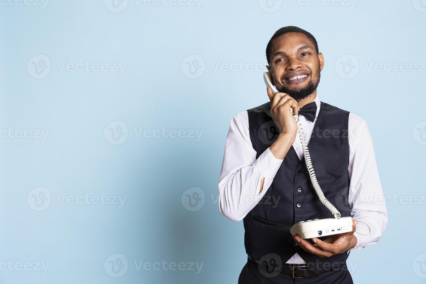 African american hotel concierge offers crucial lodging information via landline phone call, discussing about a room reservation with guests. Bellhop answering questions remotely. photo