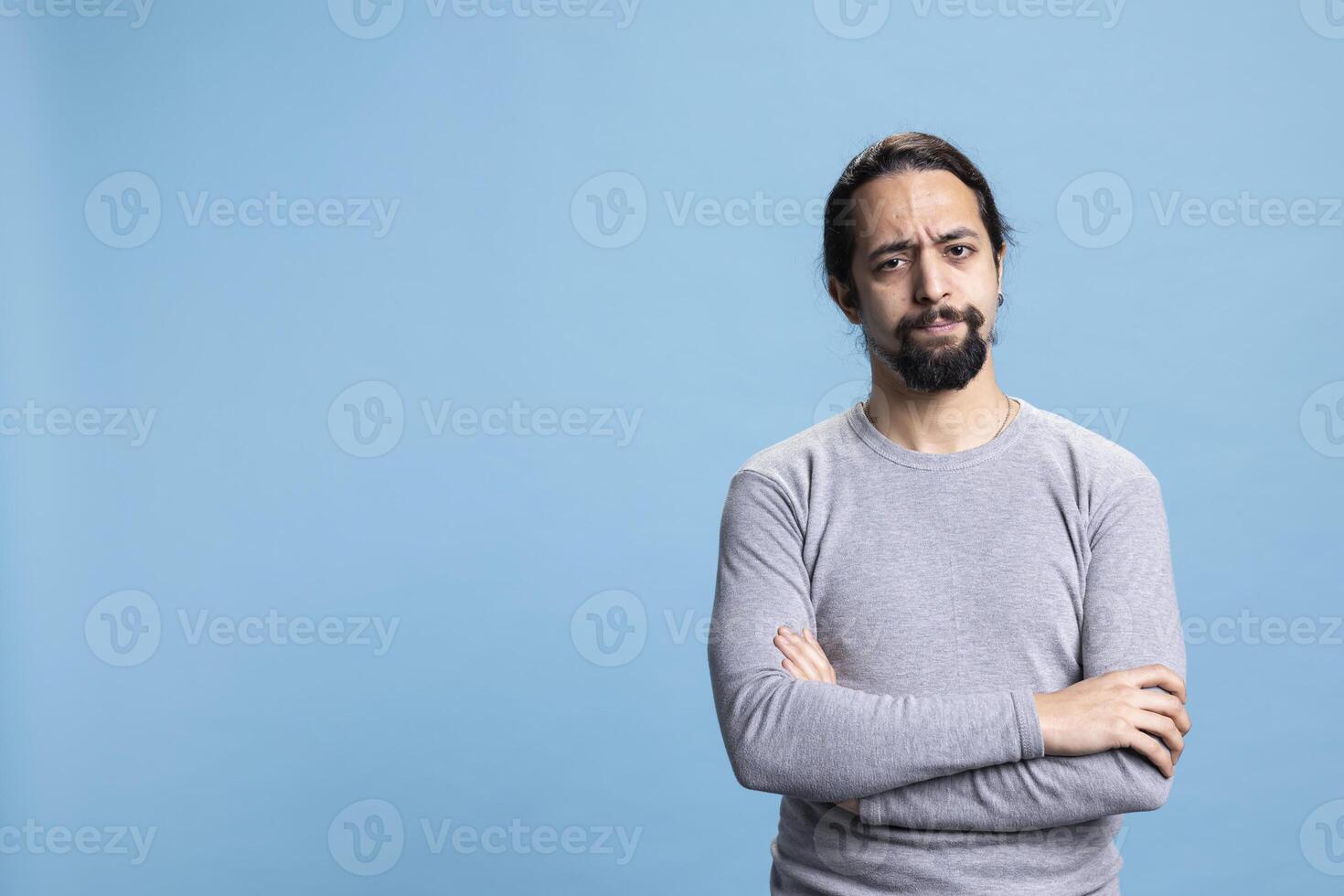 Serious person doing disappointing facial expressions on camera, pondering and posing with arms crossed against blue background. Male model with a natural authentic attitude in studio. photo