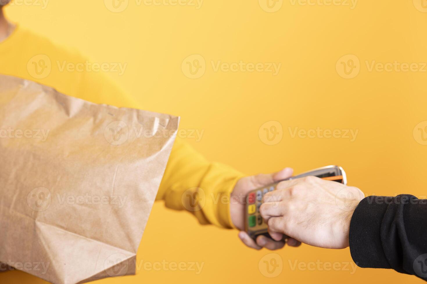 Client paying with credit card to part time delivery worker holding lunch meal order. Close up shot of man purchasing order in paper bag from courier, swiping card on POS device, studio background photo