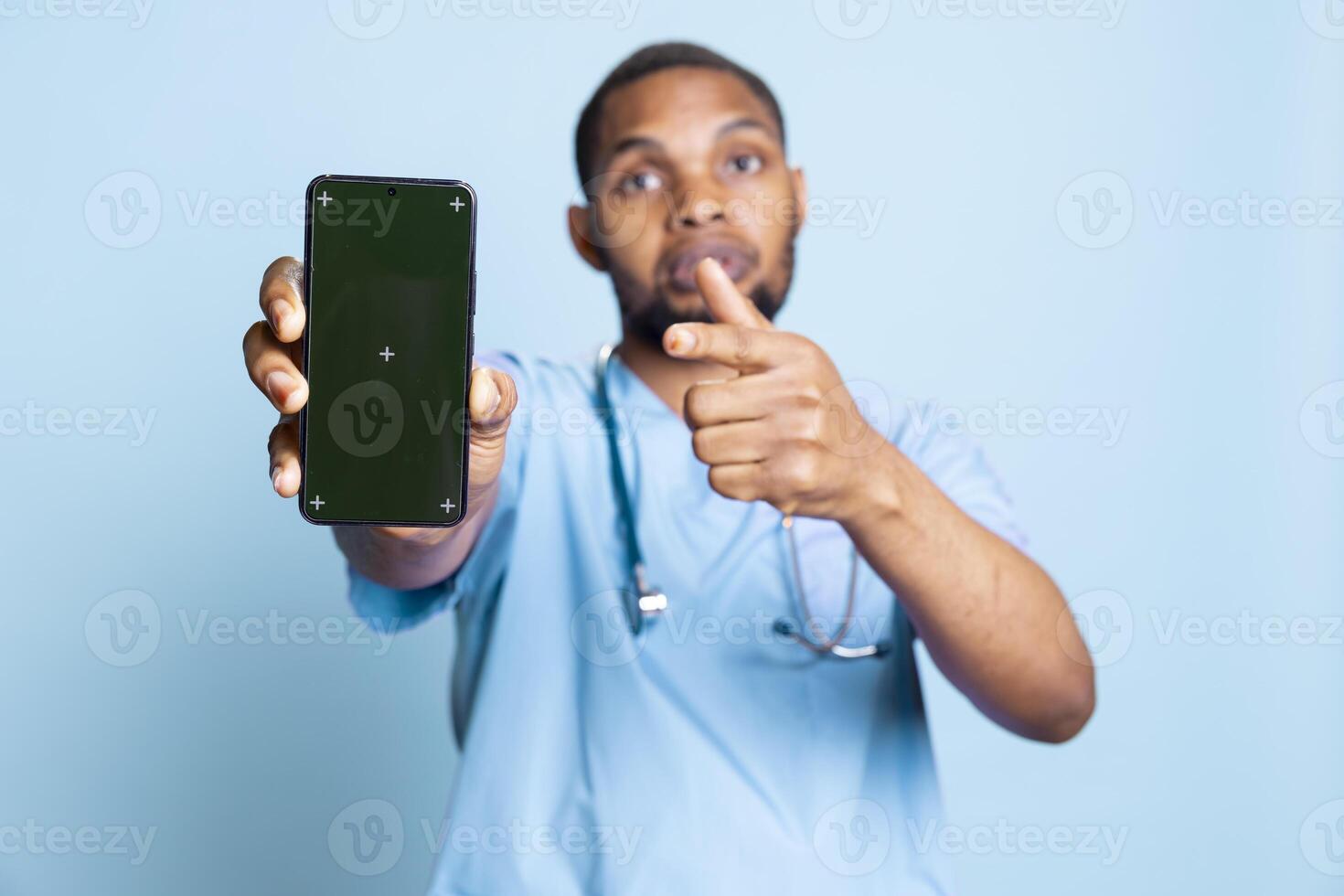 Portrait of upbeat male nurse pointing finger at green screen on smartphone. Confident hospital employee holding device with isolated chroma key display against blue studio background. photo