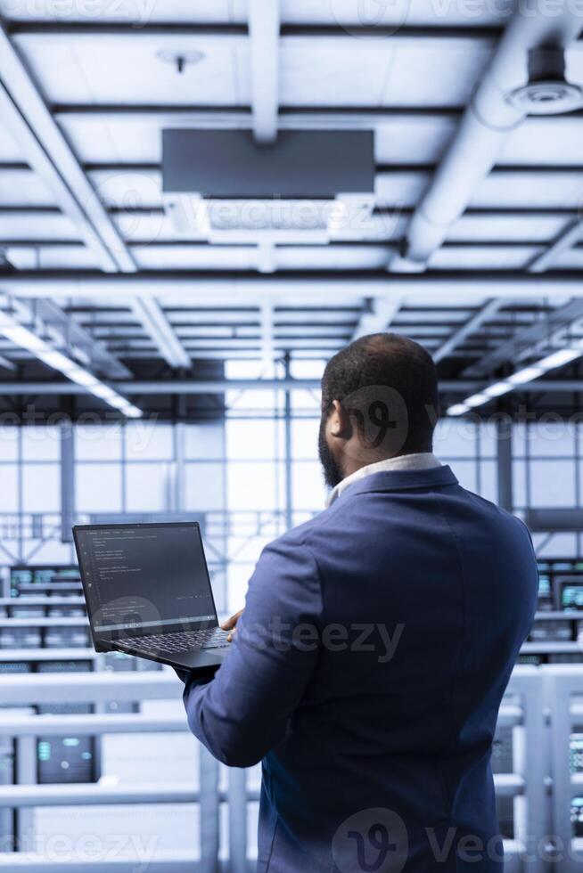 Admin in data center checking rigs and hardware, doing checkup. African american man planning and executing high tech server hub facility migrations and upgrades using notebook photo