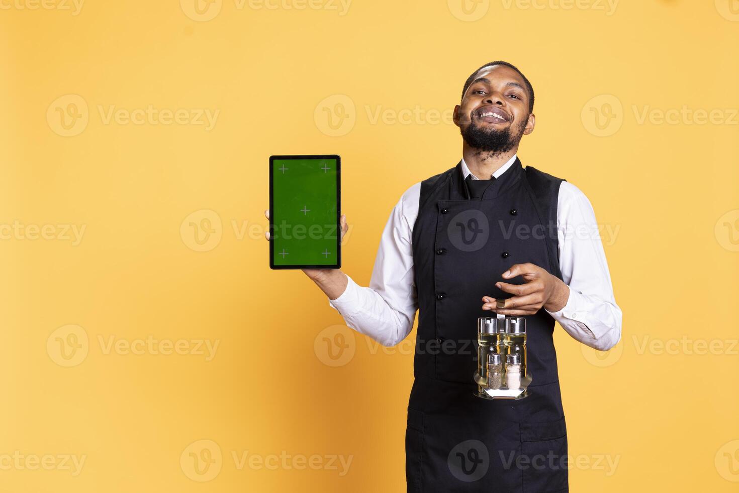 Caterer with apron and cruet set shows green screen on tablet, working in catering customer focused industry. Professional butler in uniform holds a gadget with isolated display. photo