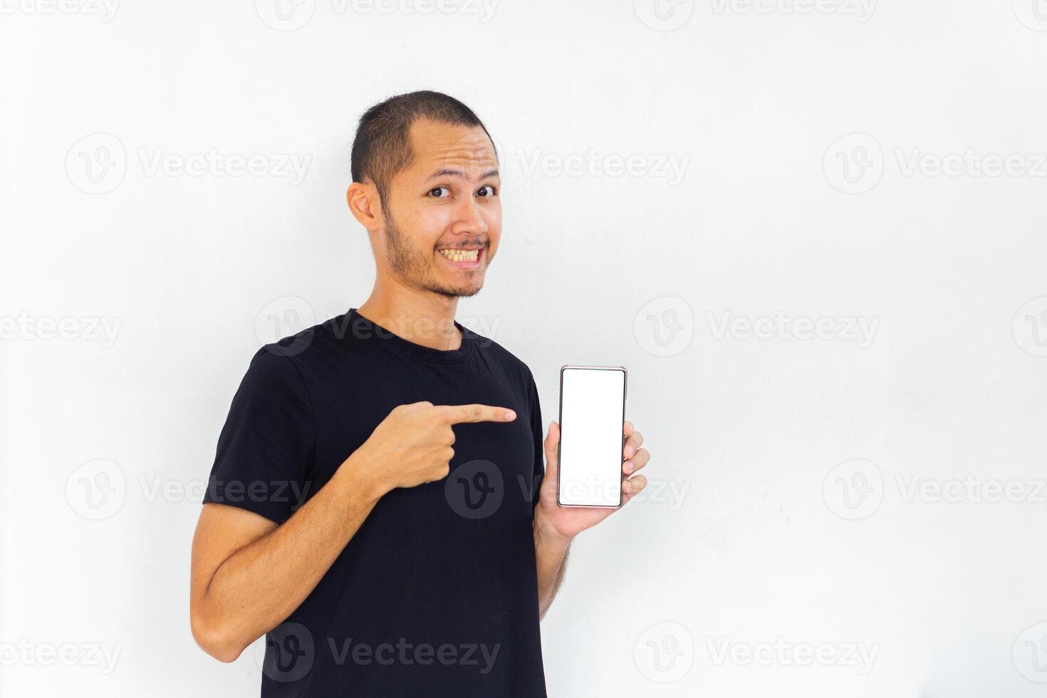 Young men with black empty shirt smiling and pointing to phone with white screen template mockup photo