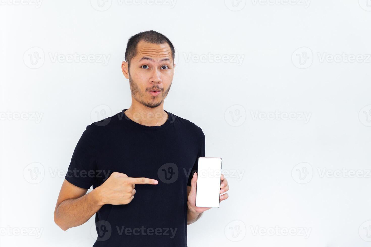 Young men with black empty shirt smiling and pointing to phone with white screen template mockup photo