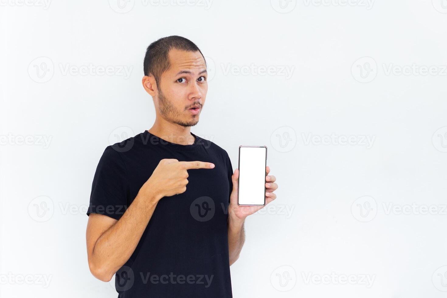 Young men with black empty shirt smiling and pointing to phone with white screen template mockup photo