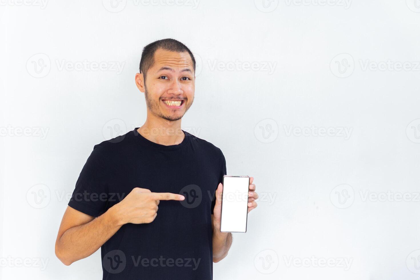 Young men with black empty shirt smiling and pointing to phone with white screen template mockup photo