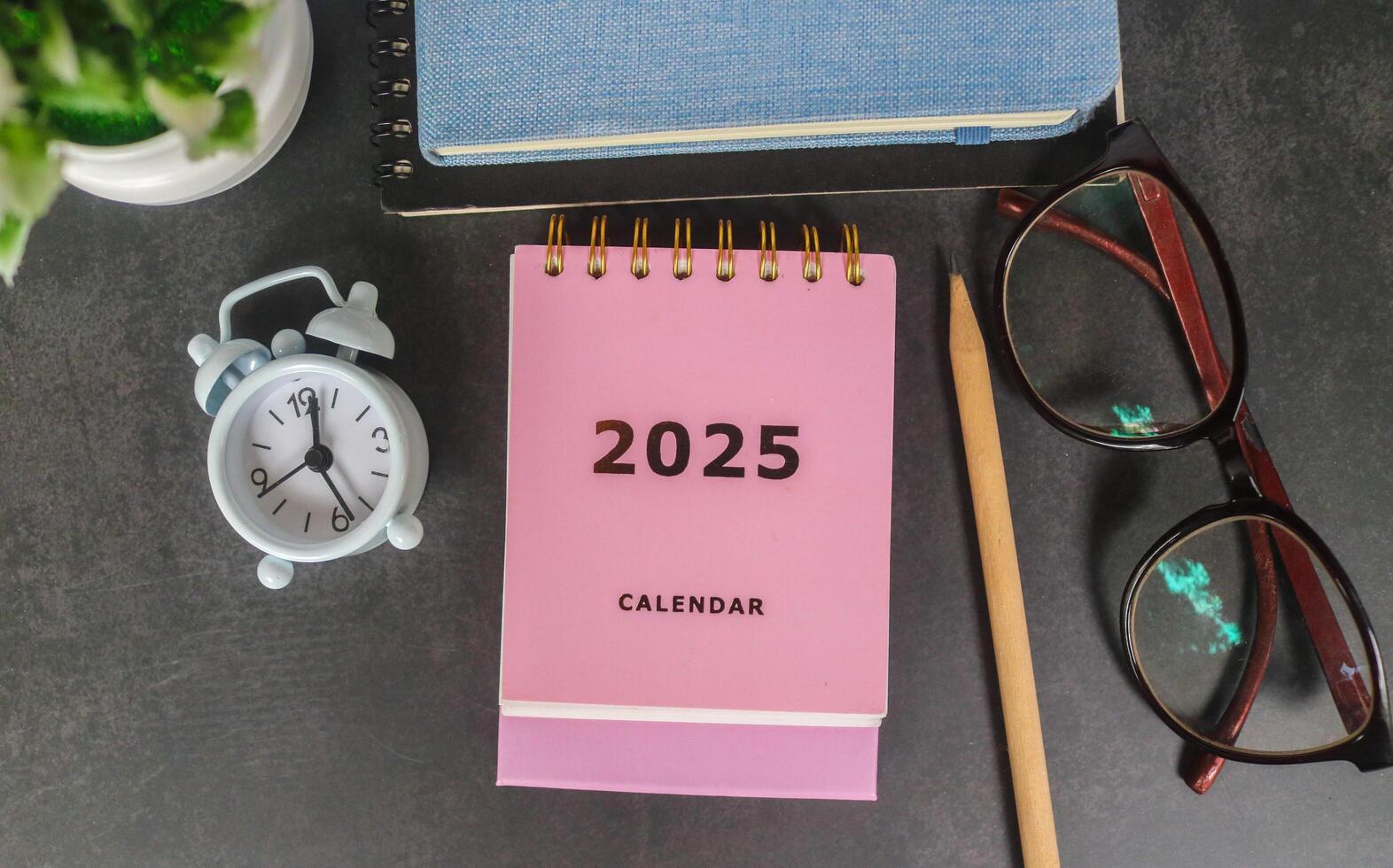 Pink calendar 2025 on office desk with notebook, glasses, pen and alarm clock. New year 2025 with office desk concept photo